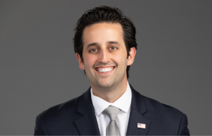 Smiling man in a dark suit, white shirt, and gray tie, wearing a small American flag pin, posed against a plain gray background—embodying professionalism and Leadership Insights.
