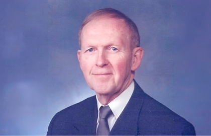 A middle-aged man with short light hair wearing a suit, dress shirt, and tie poses for a formal studio portrait against a blue background, reflecting professionalism and Leadership Insights.