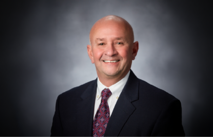 A bald man wearing a dark suit, white shirt, and patterned tie smiles in a professional studio portrait with a gray background, radiating confidence and ready to share Leadership Insights.