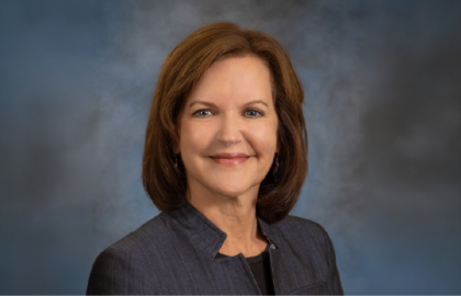 A woman with shoulder-length brown hair, wearing a dark blazer, poses confidently against a blue and gray studio background, embodying Leadership Insights.