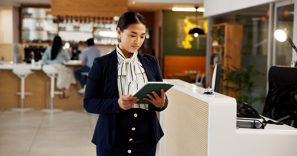 A woman in business attire stands at a reception desk, looking at a tablet in a modern office or hotel lobby, suggesting the efficiency and organization often seen in health care management settings.