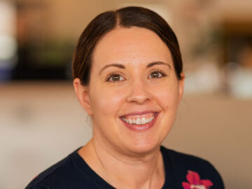 A woman with brown hair pulled back, wearing a navy sweater with a floral pattern, smiles at the camera in an indoor setting, celebrating her recognition as a 20 Under 40 Alumni.