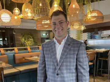 A 20 Under 40 Alumni wearing glasses, a plaid blazer, white shirt, and dark pants stands and smiles in a modern restaurant with wooden tables and woven hanging lights.