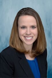 A 20 Under 40 alumni with straight, shoulder-length brown hair, wearing a black blazer over a blue top, smiles at the camera against a light blue mottled background.