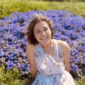 A 20 Under 40 alumni, a young woman in a light floral dress, sits on grass before a large patch of vibrant purple flowers, smiling at the camera.
