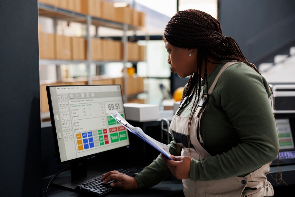 A woman wearing overalls reviews paperwork while using a desktop computer with a point-of-sale system, illustrating day-to-day operations and supply chain management in a warehouse or industrial workspace.