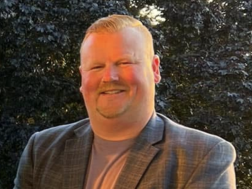 A 20 Under 40 alumni in a gray plaid blazer, light purple shirt, and jeans stands outdoors by a metal railing, with green grass and trees in the background.