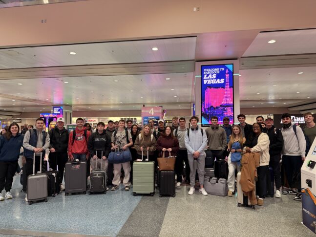 A group of people with luggage poses for a photo in an airport terminal near a "Welcome to Fabulous Las Vegas" sign.