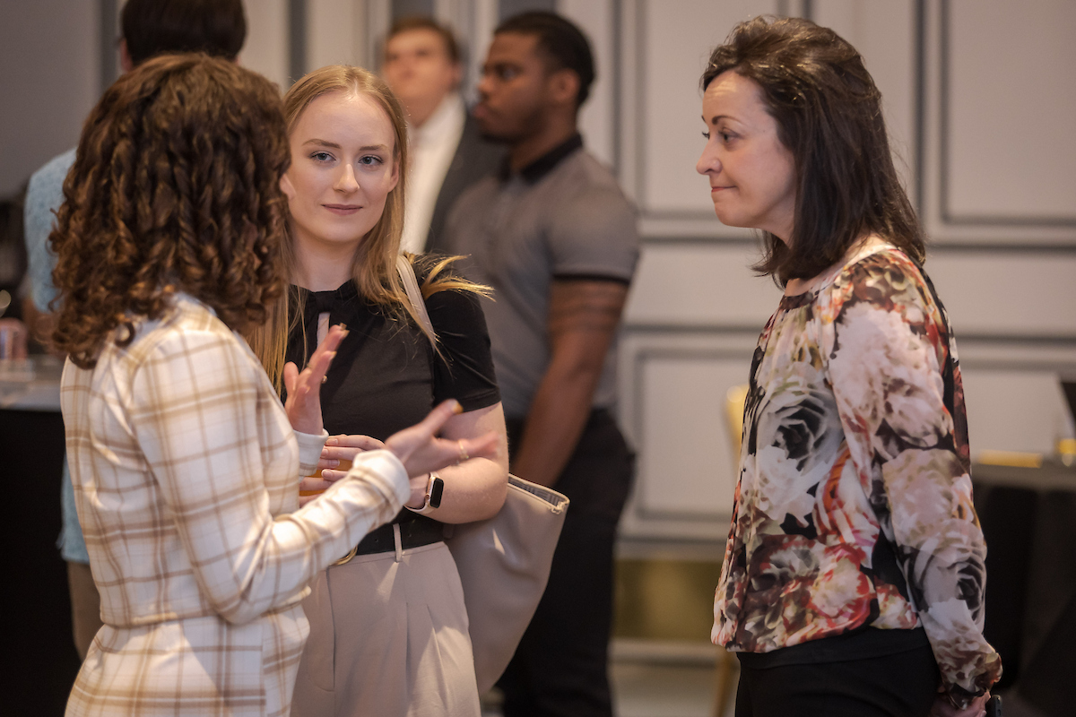 Three women are having a conversation about health care management indoors while other people stand in the background.