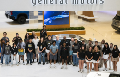 A group of young people poses for a photo under a large "general motors" sign inside an indoor showroom with vehicles in the background.