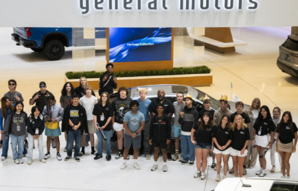 A group of young people poses for a photo under a large "general motors" sign inside an indoor showroom with vehicles in the background.