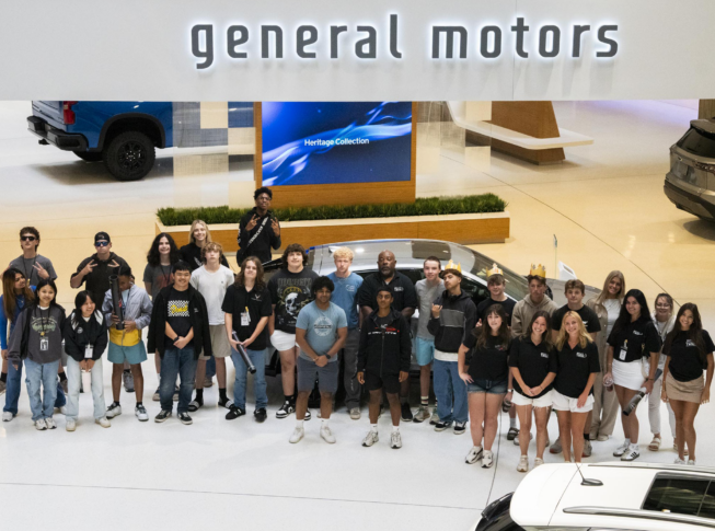 A group of young people poses for a photo under a large "general motors" sign inside an indoor showroom with vehicles in the background.