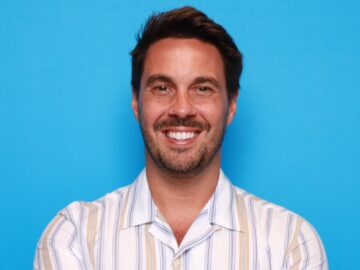 Alumni and 20 Under 40 honoree, this man with short dark hair, beard, and mustache smiles with arms crossed in a striped short-sleeved shirt, standing confidently before a plain blue background.