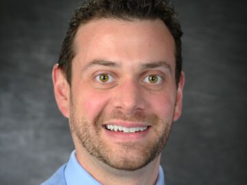 A man with short brown hair and a trimmed beard, wearing a light blue suit jacket, blue shirt, and patterned tie, poses in front of a dark gray background—an accomplished Alumni and member of the 20 Under 40.