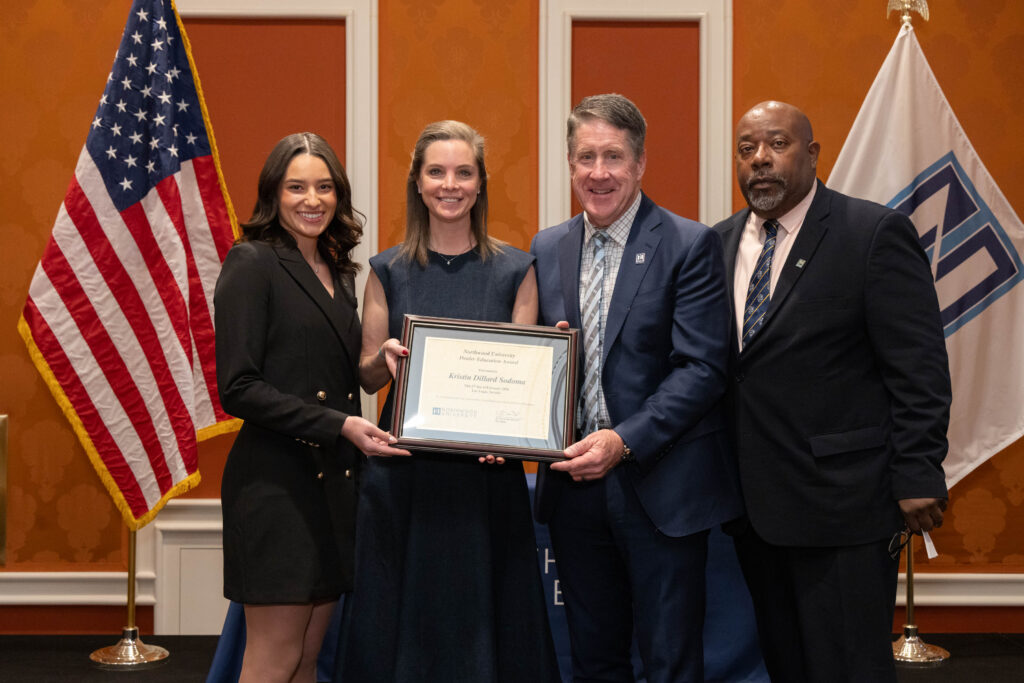Four people in formal attire stand indoors; two women hold a framed certificate. U.S. and Northwood University flags are displayed in the background, highlighting excellence among automotive leaders.