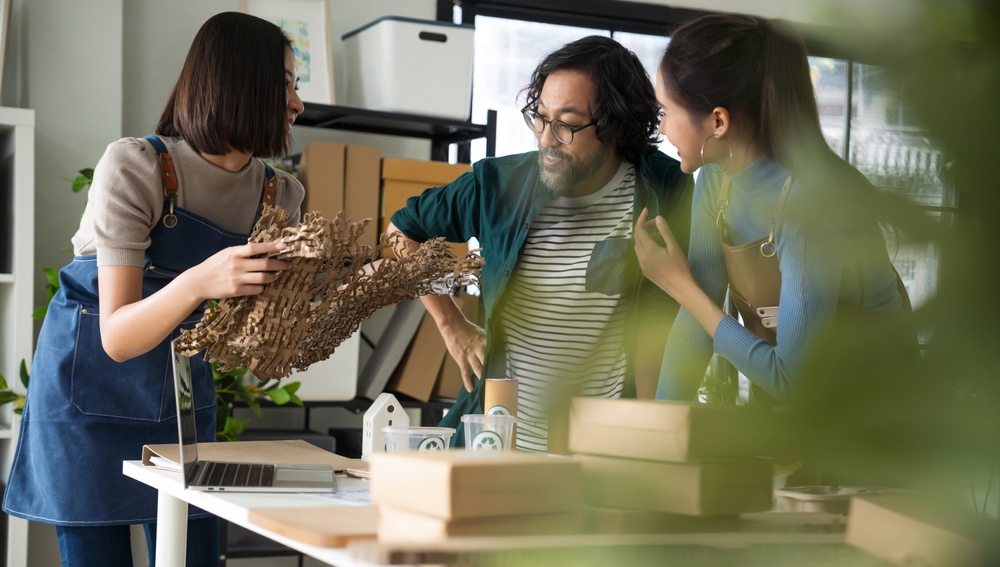 Three people in aprons examine a wooden or cardboard sculptural piece in a modern workspace, discussing ideas that could inspire innovative designs for health care management environments. Boxes, laptops, and model houses fill the table.
