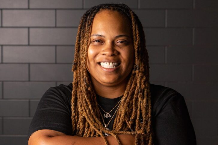 A person with long, light brown dreadlocks and a black t-shirt smiles in front of a dark tiled wall.