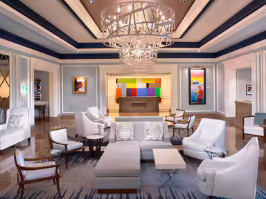 A spacious hotel lobby with modern white furniture, a large chandelier, colorful artwork on the walls, and polished floors—an inviting space perfect for Alumni gatherings or celebrating 20 Under 40 honorees.