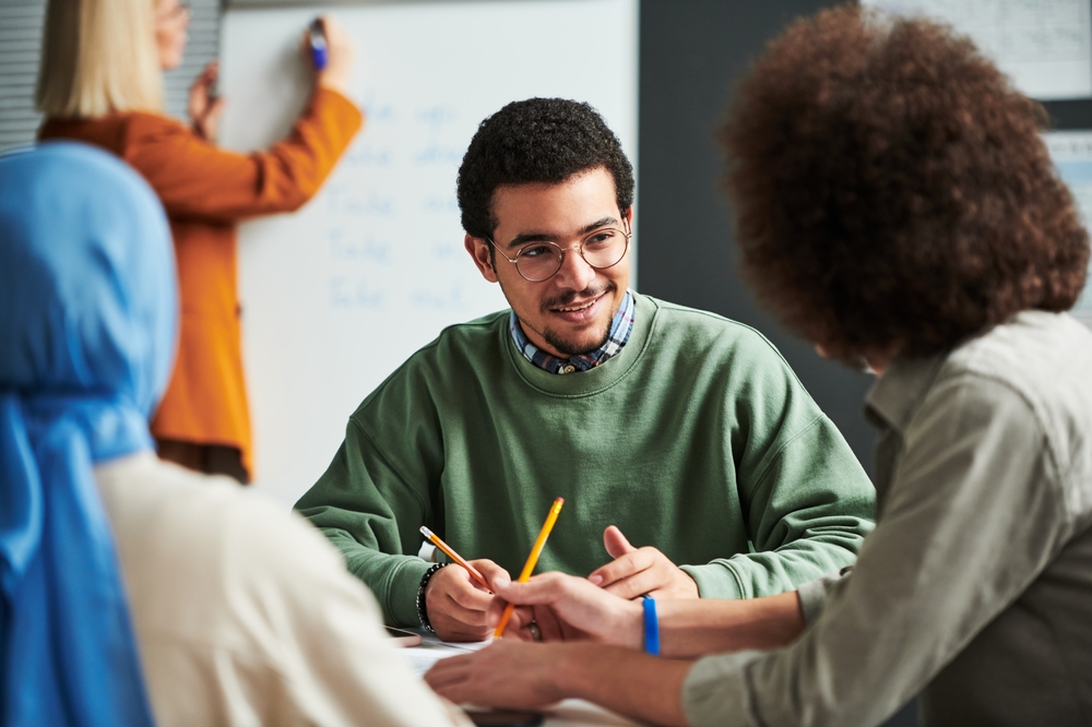 Four people are in a meeting room discussing international business. Three sit around a table talking and taking notes, while one stands at a whiteboard writing.