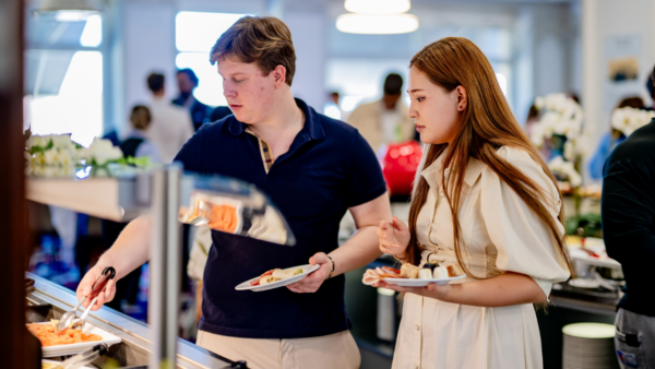 Two people serve themselves food from a buffet table during an Undergraduate Admissions event, each holding a plate and choosing items, with other diners and food trays visible in the background.