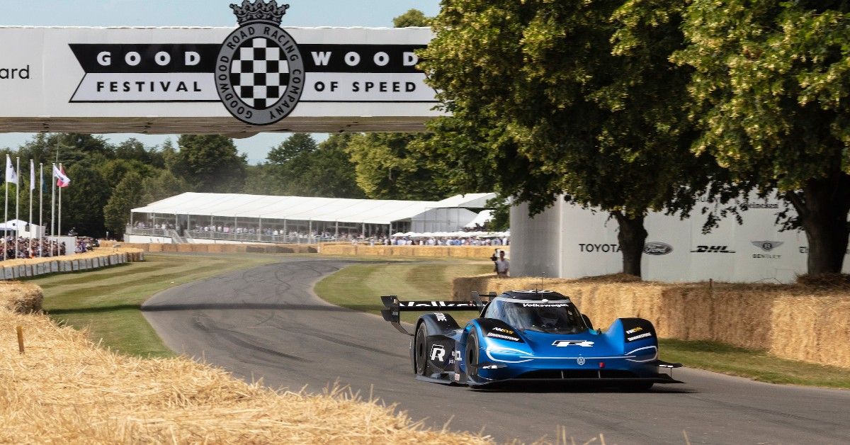 A blue electric race car drives on a track at the Goodwood Festival of Speed, perfect for a Family Trip, passing under a banner with trees and spectator tents in the background.
