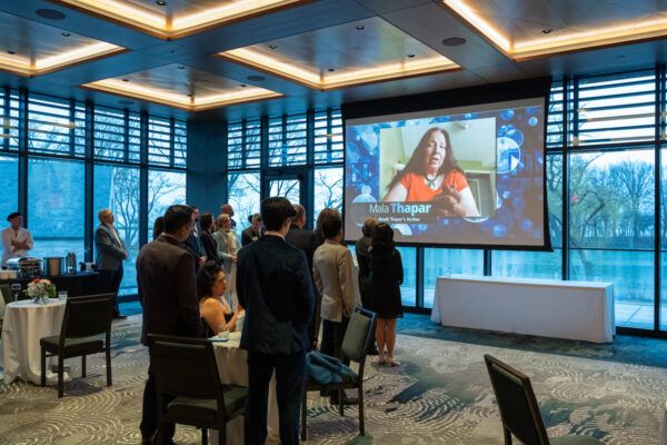 A group of people in formal attire watch a large screen displaying a woman speaking at the Outstanding Business Leader Awards in a modern, windowed event space.