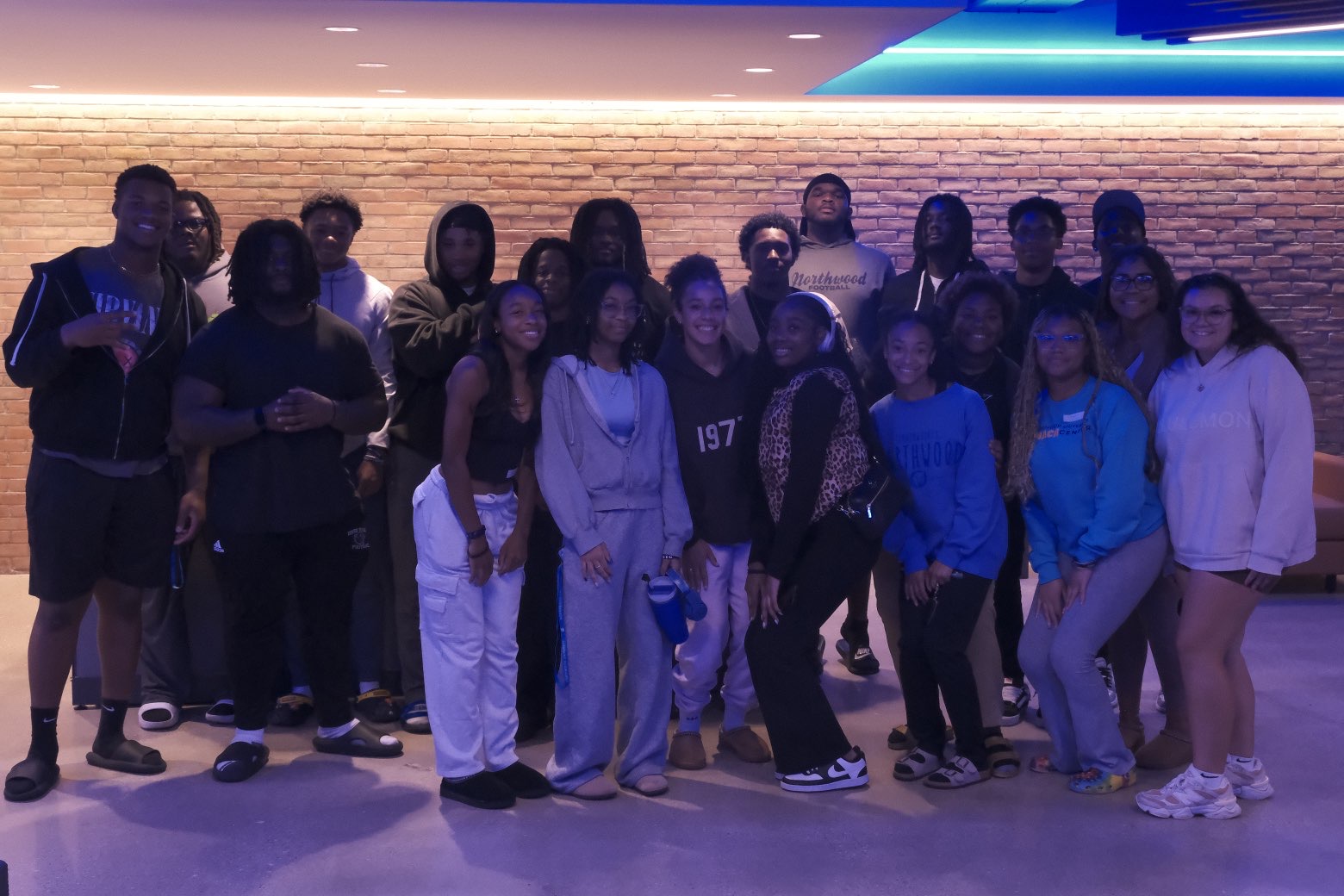 A group of people from the Black Student Union pose together indoors in front of a brick wall, dressed in casual attire and displaying relaxed expressions under blue ceiling lights.