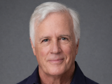 An older man with short white hair wearing a dark blue collared shirt, smiling slightly, poses in front of a plain gray background, embodying the confidence of an Outstanding Business Leader Awards recipient.