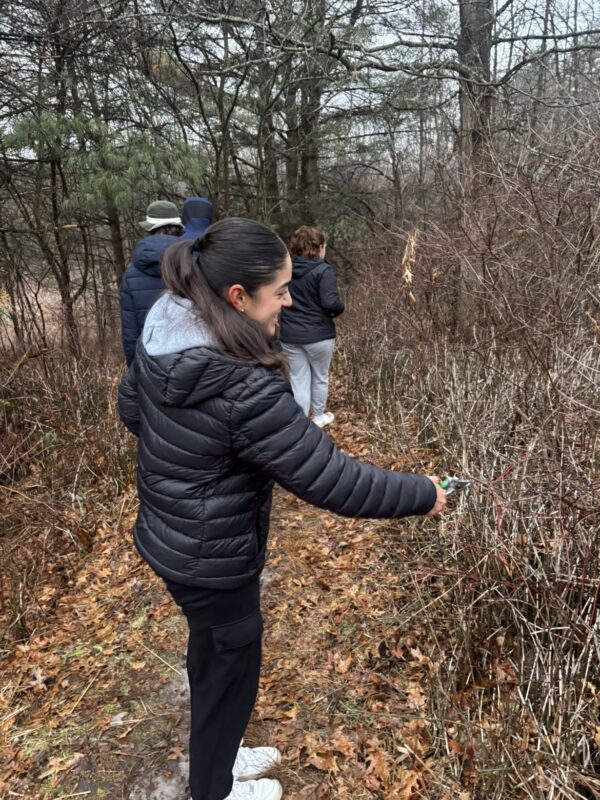 Members of the Student Honors Association, dressed in jackets and sneakers, walk along a leaf-covered forest trail on a cloudy day. One person in the foreground is holding a pair of pruning shears.