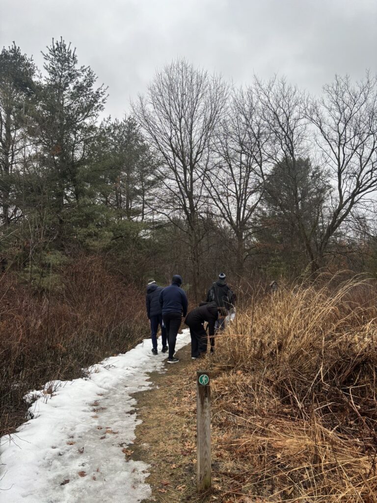 Four members of the Student Honors Association walk on a partially snow-covered trail through a wintery landscape with bare trees and tall grass under a cloudy sky.