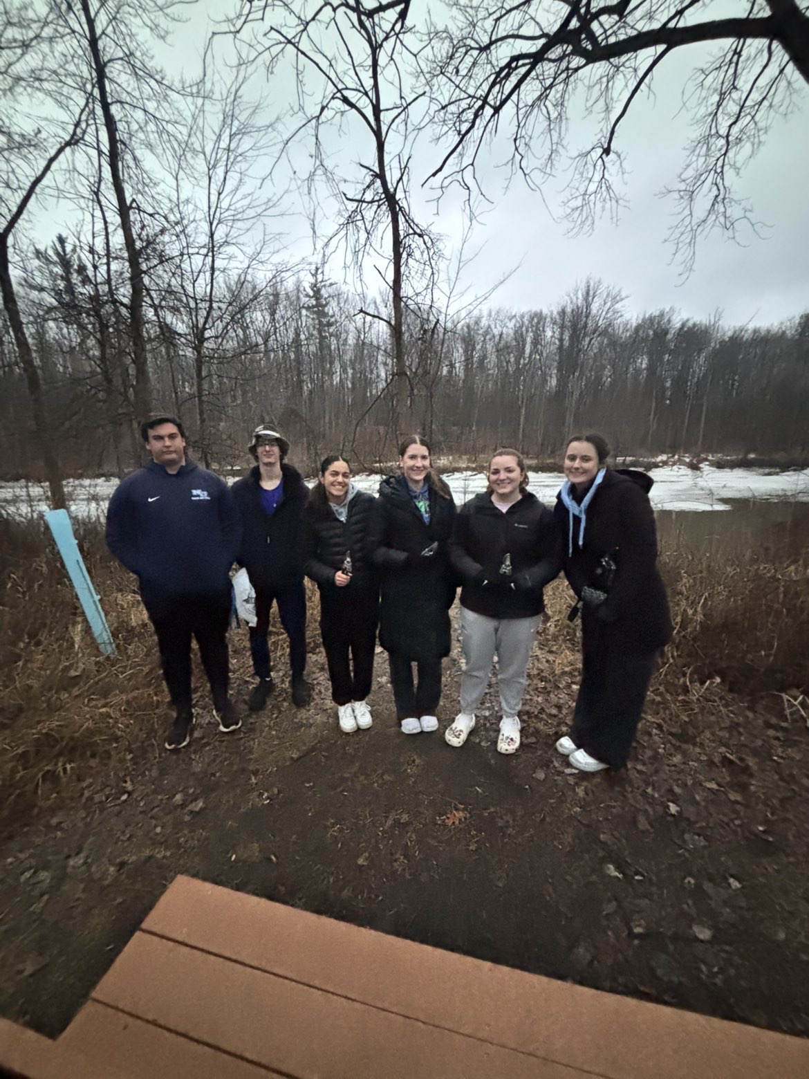 Six members of the Student Honors Association stand outdoors on a dirt path near a body of water, with leafless trees in the background under an overcast sky.