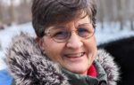 A woman with short gray hair and glasses smiles while wearing a green coat with a fur-lined hood. Snow and trees are visible in the blurred background.