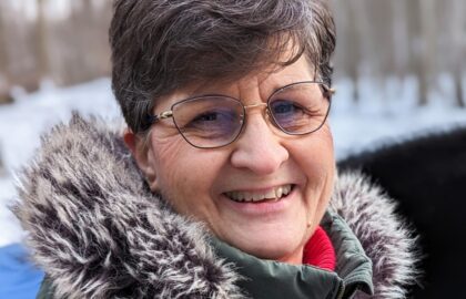 A woman with short gray hair and glasses smiles while wearing a green coat with a fur-lined hood. Snow and trees are visible in the blurred background.
