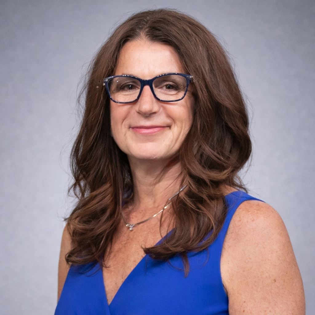 A woman with long brown hair, wearing glasses and a blue sleeveless top, smiles at the camera against a plain gray background.