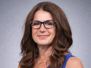A woman with long brown hair and glasses, wearing a blue sleeveless top, stands in front of a plain gray background, smiling slightly at the cameraโcapturing the poised confidence of an Outstanding Business Leader Awards recipient.