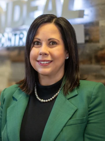 A woman with straight dark hair, wearing a green blazer, black top, pearl necklace, and stud earrings, poses indoors with a wooden wall and a blurred Northwood University Outstanding Business Leaders 2026 sign in the background.