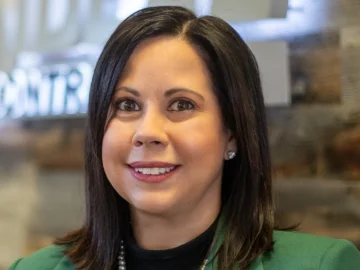 A woman with straight dark hair, wearing a green blazer, black top, pearl necklace, and stud earrings, stands indoors in front of a blurred Outstanding Business Leader Awards sign and wood-paneled wall.