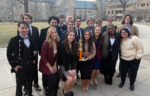 A group of young adults in formal attire poses outdoors on a paved path, with several holding a gold trophy; buildings and leafless trees are in the background.