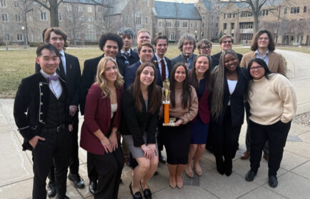 A group of young adults in formal attire poses outdoors on a paved path, with several holding a gold trophy; buildings and leafless trees are in the background.