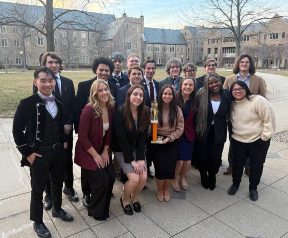 A group of young adults in formal attire poses outdoors on a paved path, with several holding a gold trophy; buildings and leafless trees are in the background.
