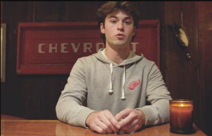 A young man in a grey hoodie with a red wing emblem sits at a wooden table in front of a red Chevrolet sign, with a lit candle to his side.
