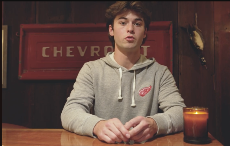 A young man in a grey hoodie with a red wing emblem sits at a wooden table in front of a red Chevrolet sign, with a lit candle to his side.