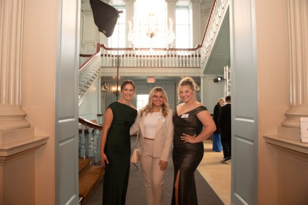 Three women from the Signature Events Team, dressed in formal attire, stand smiling in the entrance of an elegant hall with a chandelier and staircase in the background.