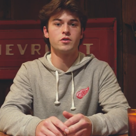 A young man in a gray hoodie with a red winged wheel logo sits at a wooden table, a candle lighting his thoughtful face, with a red Chevrolet sign glowing in the background—capturing the quiet hope of the future.