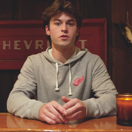 A young man in a gray hoodie with a red winged wheel logo sits at a wooden table, a candle lighting his thoughtful face, with a red Chevrolet sign glowing in the background—capturing the quiet hope of the future.
