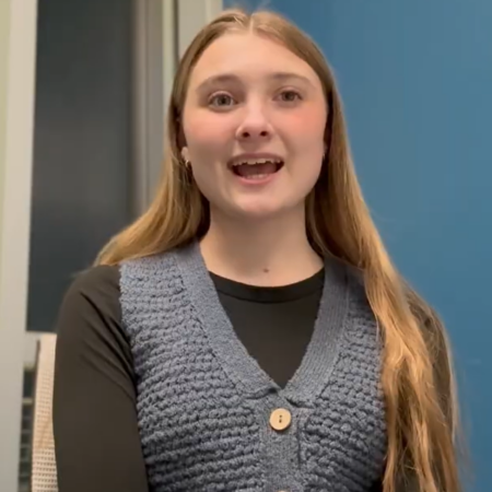 A young woman with long hair sits indoors in front of a blue wall, wearing a black shirt and a blue knit vest, looking slightly upward with a neutral expression as soft lights hint at dreams of the future.