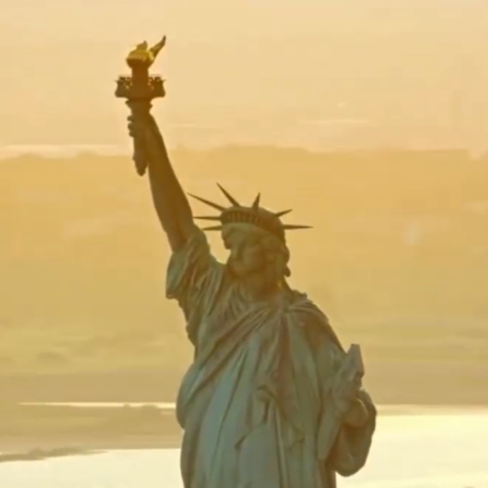 The Statue of Liberty stands with torch raised, shining as a beacon of freedom against a hazy, golden sky, with water and distant land evoking hope for the future.