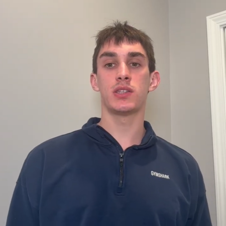 A young man in a dark blue Gymshark sweatshirt stands indoors near a light gray wall and a white door, quietly reflecting on his future under the soft lights.