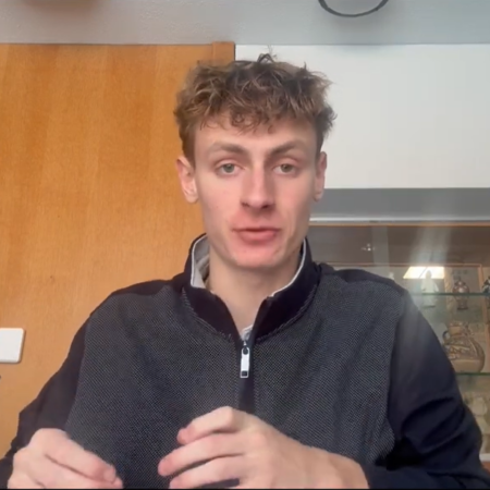 A young man with light brown hair sits at a table indoors, wearing a dark zip-up jacket, as warm wooden walls and a glass display case frame the scene—capturing a quiet moment where freedom lights the future.