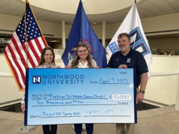 Three people stand indoors holding a large ceremonial check for ,000 from Northwood University’s Project 100, dated April 19, 2025, with U.S. and state flags in the background.